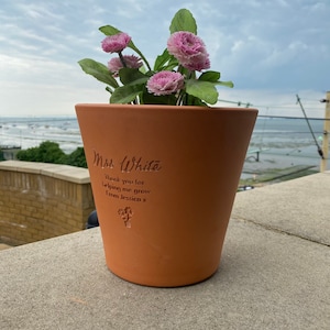 May include: A terracotta flower pot with a personalised message that reads "Mrs. White Thank you for helping me grow From Jessica x". The pot is filled with pink flowers and is sitting on a concrete surface.