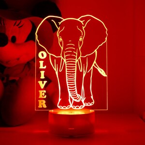 May include: A glowing, personalized night light featuring an elephant design. The light is a clear acrylic panel with the name "OLIVER" vertically displayed. The elephant is illuminated in a warm, inviting glow, resting on a round base.