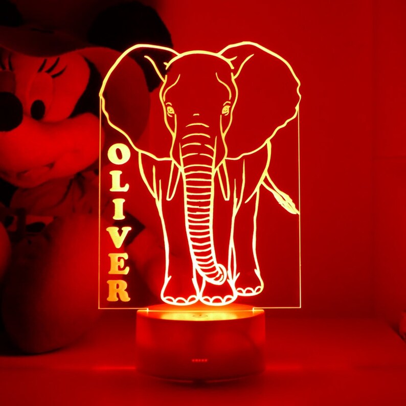 May include: A glowing, personalized night light featuring an elephant design. The light is a clear acrylic panel with the name "OLIVER" vertically displayed. The elephant is illuminated in a warm, inviting glow, resting on a round base.