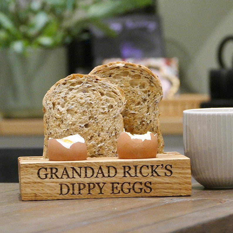 May include: Wooden dippy egg holder with two soft boiled eggs and two slices of bread. The holder is engraved with the text "Grandad Rick's Dippy Eggs".