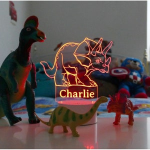 May include: A red light-up dinosaur nightlight with a white base and the name "Charlie" printed on the front. The nightlight is surrounded by three toy dinosaurs.