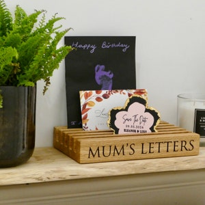 May include: Wooden letter holder with the text "MUM'S LETTERS" engraved on the front. The holder has slots for cards and envelopes. There are three cards in the holder, one with the text "Happy Birthday", one with a floral design, and one with a pink flower and the text "Save The Date 09.05.2024 ELEANOR & LUKR".