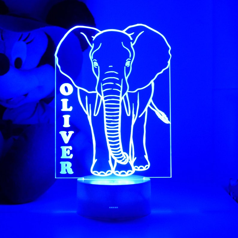 May include: A blue LED night light featuring a white outline of an elephant. The name "OLIVER" is vertically displayed on the left side of the elephant. The light sits on a dark base, creating a glowing effect.
