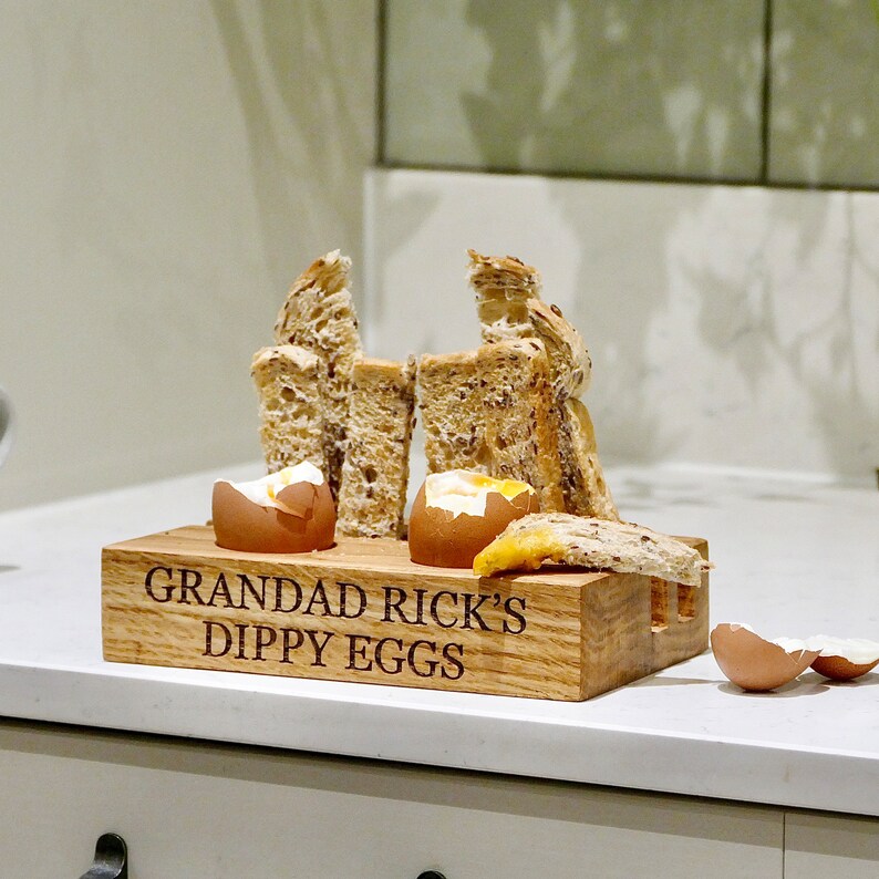 May include: A wooden serving tray with the text "Grandad Rick's Dippy Eggs" engraved on it. The tray is filled with two soft-boiled eggs and several slices of toasted bread.