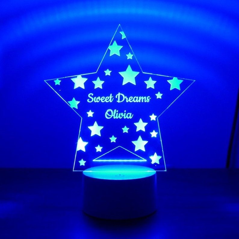 May include: A white star-shaped night light with the words "Sweet Dreams Olivia" inscribed on it. The star is surrounded by smaller stars and is lit up with a blue light.