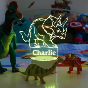 May include: A white night light shaped like a triceratops dinosaur with the name "Charlie" printed below. The light is on and glowing green. There are three toy dinosaurs in the image.