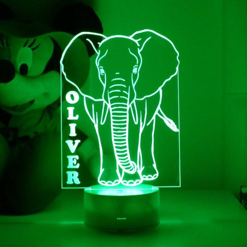 May include: A green LED night light featuring a white outline of an elephant. The name "OLIVER" is vertically displayed on the left side. The lamp sits on a dark green base, illuminating the elephant design. A Minnie Mouse plush toy is in the background.