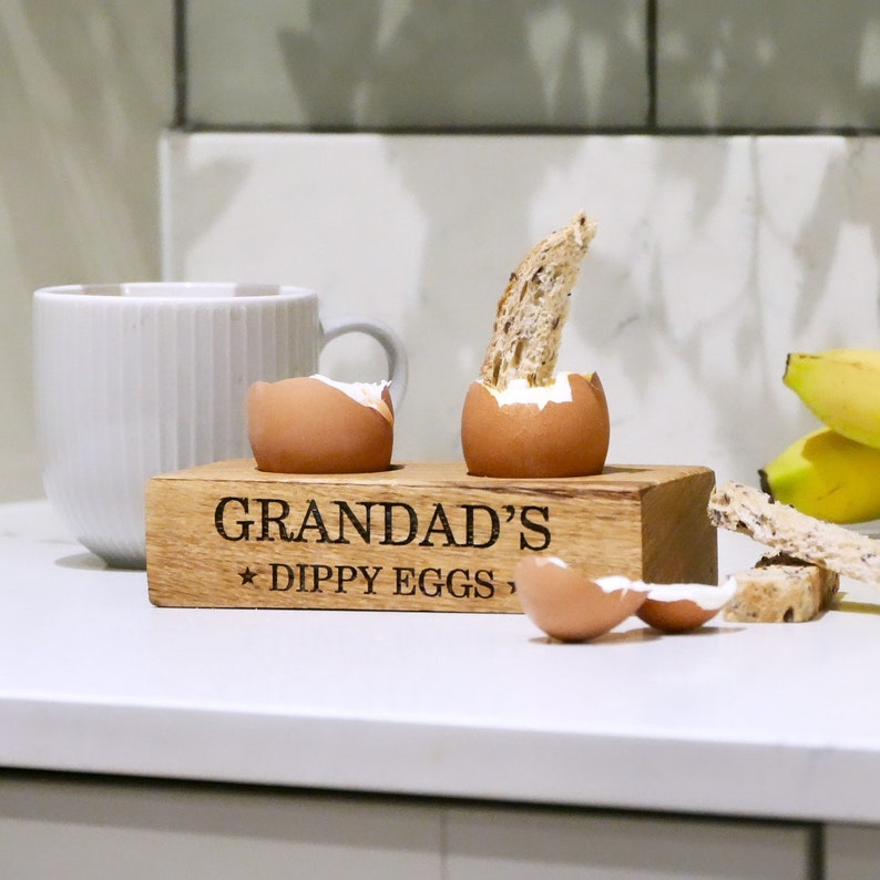 May include: Wooden egg holder with the text "Grandad's Dippy Eggs" engraved on the front. Two eggs are in the holder, one with a piece of toast in it.