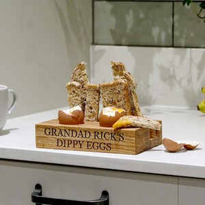 May include: A wooden egg holder with the text "Grandad Rick's Dippy Eggs" engraved on the front. Two boiled eggs are in the holder, and there are pieces of bread beside them.