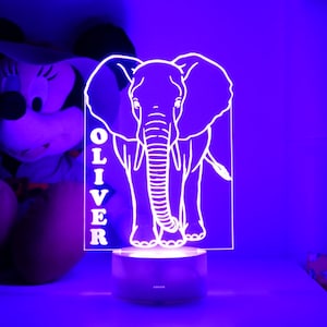 May include: A white elephant-shaped night light with the name "OLIVER" vertically displayed. The light sits on a dark base, emitting a soft glow. The design is a modern, personalized touch for a child's room.