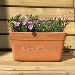 May include: Terracotta planter with the text "Mr & Mrs Lowdell Est 23.04.2022" engraved on the front. The planter is filled with pink flowers.
