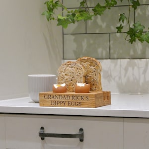 May include: Wooden egg holder with two soft boiled eggs and two slices of bread. The holder is engraved with the text "Grandad Rick's Dippy Eggs".