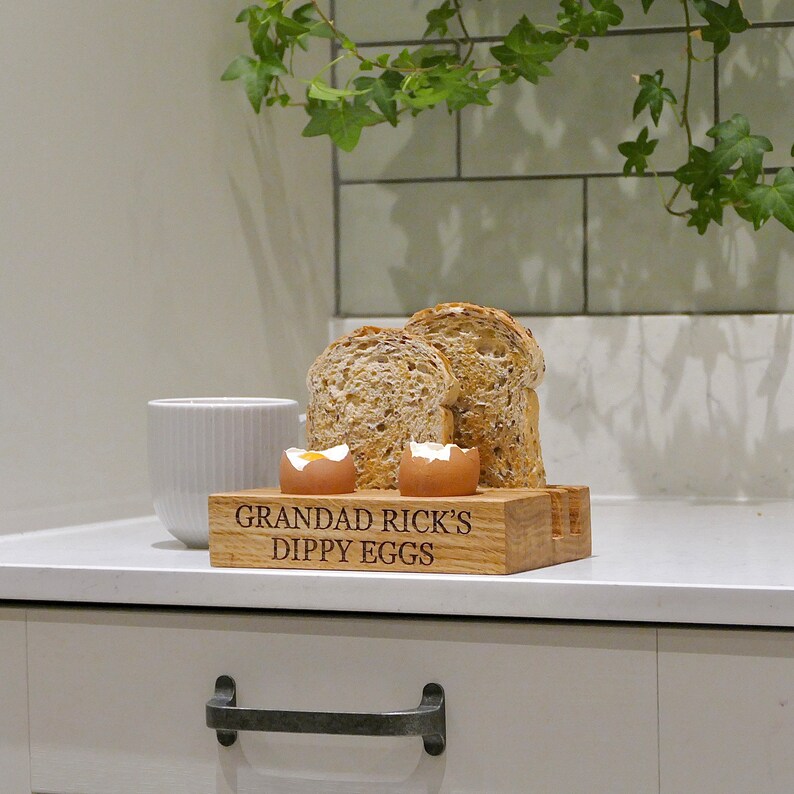 May include: Wooden egg holder with two soft boiled eggs and two slices of bread. The holder is engraved with the text "Grandad Rick's Dippy Eggs".