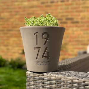 May include: A grey concrete planter with the year 1974 engraved on the front. The planter has the text "Happy 50th Dad Love Jess & Sam" engraved below the year.