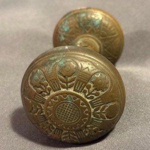 May include: Two antique brass doorknobs with floral and geometric patterns. The knobs have a weathered patina, showing signs of age and use. The design includes a central sunburst motif surrounded by stylized flowers and leaves.