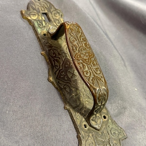 May include: Antique bronze door handle with ornate floral and scrollwork detailing. The handle features a curved grip and a rectangular backplate with mounting holes. The metal has a weathered patina, suggesting age and use. A decorative piece for vintage home decor.