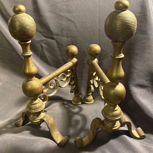 May include: A pair of antique brass andirons. Each andiron features a decorative, ribbed sphere finial atop a slender, curved post. The andirons have ornate, scrolled crossbars and curved feet, showcasing an aged patina.