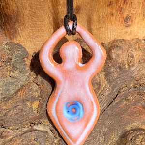 May include: A ceramic pendant shaped like a female figure with raised arms. The pendant is painted in shades of pink, white and blue, with a blue and white swirl design in the centre.
