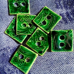 May include: A collection of square, glossy, emerald green ceramic buttons. Each button features two or three holes for sewing and a decorative swirl pattern. The buttons are scattered on a textured blue surface.