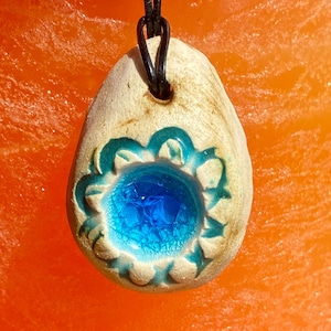 May include: A white ceramic pendant with a blue glass center and a floral design. The pendant is on a black leather cord.