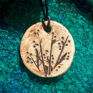 Puede incluir: Un colgante redondo de cerámica con un cordón de cuero negro. El colgante presenta un diseño de flores estilizadas con tallos marrones y puntos blancos.