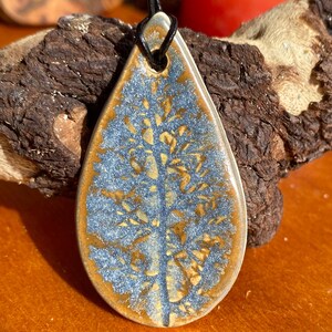 May include: A teardrop-shaped ceramic pendant featuring a glossy finish with a blue and tan leaf-like pattern. The pendant has a hole at the top for a black cord, showcasing a detailed botanical impression with a central stem and branching veins.
