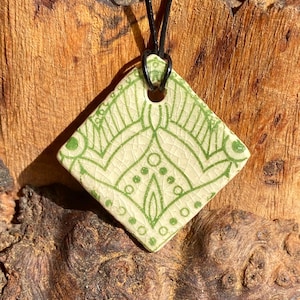 Puede incluir: Un colgante cuadrado de cerámica con un diseño geométrico verde y blanco, colgado de un cordón de cuero negro.