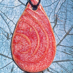 May include: A teardrop-shaped pendant necklace made of red and white ceramic with a swirling pattern. The pendant is hanging from a black cord.