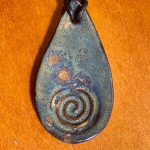 May include: A teardrop-shaped ceramic pendant featuring a mottled blue and brown glaze with a spiral design embossed in the center. The pendant is suspended from a dark leather cord that passes through a hole at the top of the ceramic piece.