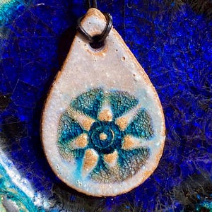 May include: A teardrop-shaped ceramic pendant with a blue and gold floral design. The pendant has a crackled glaze and is strung on a black cord.