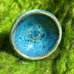 May include: A small, round, blue and gold ceramic bowl with a speckled glaze. The bowl is sitting on a bed of green moss.