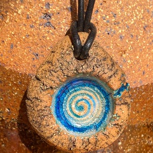 Puede incluir: Un colgante en forma de lágrima hecho de arcilla marrón texturizada con una incrustación circular central con un patrón de espiral azul y blanco. El colgante cuelga de un cordón de cuero negro que pasa por un lazo en la parte superior.