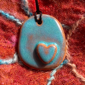 May include: A turquoise ceramic pendant with a smaller heart-shaped pendant attached. The pendant is on a black cord.