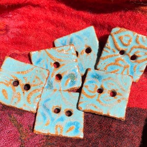 May include: Five square ceramic buttons with a blue glaze and gold flecks. The buttons have a swirling pattern and two holes each.