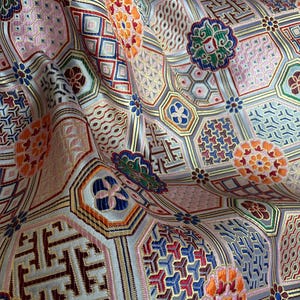 May include: Detailed shot of a patterned fabric with a complex design. The fabric features a repeating geometric pattern with various shapes, including hexagons and floral motifs. Colours include pink, orange, blue, green, and gold, creating a rich textile.