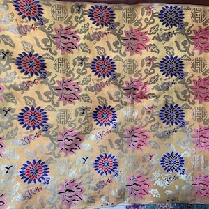 May include: A golden-yellow textile with a repeating floral pattern. The design features pink, blue, and red flowers, along with gray and silver decorative elements. The fabric has a subtle sheen.