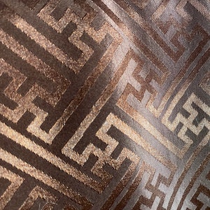 May include: Close-up of a brown fabric with a gold geometric pattern. The pattern is a repeating design of interlocking squares and lines.