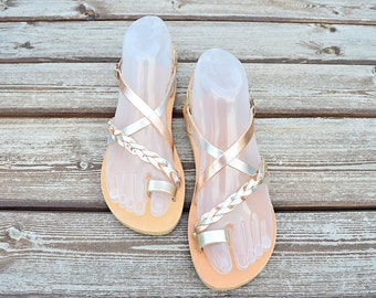 May include: A pair of gold and silver leather sandals with a braided strap design. The sandals have a toe ring and an ankle strap.