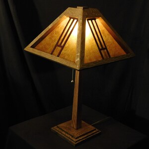 May include: A wooden table lamp with a square shade. The shade has a geometric design with vertical and horizontal lines. The lamp is made of wood and has a warm, inviting glow. The lamp is on a square base.