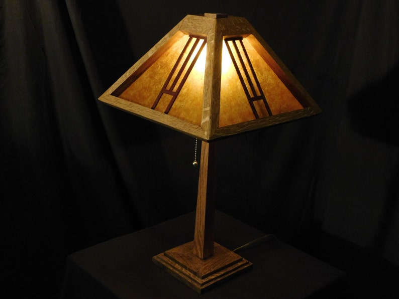 May include: A wooden table lamp with a square shade. The shade has a geometric design with vertical and horizontal lines. The lamp is made of wood and has a warm, inviting glow. The lamp is on a square base.