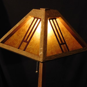 May include: A wooden Arts and Crafts style lamp with a square shade. The shade is made of light-colored wood with a warm, amber-toned paper insert. The lamp has a pull chain and a dark background.