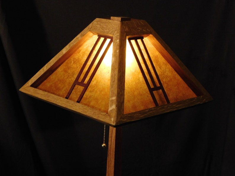 May include: A wooden Arts and Crafts style lamp with a square shade. The shade is made of light-colored wood with a warm, amber-toned paper insert. The lamp has a pull chain and a dark background.