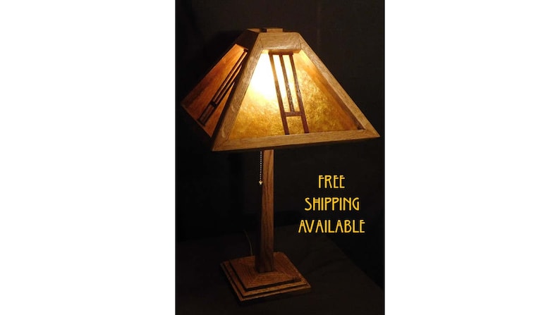 May include: A wooden table lamp with a geometric shade. The shade is a light tan color with dark brown accents. The lamp has a square base and a tall, slender stem. The text "FREE SHIPPING AVAILABLE" is visible.