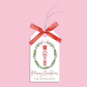 May include: A white gift tag with a red bow and a pink and red nutcracker illustration. The tag has the text "Merry Christmas" and "THE JOHNSON'S" printed on it.