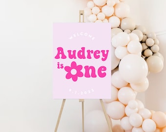 May include: A pink welcome sign on a wooden easel. The sign reads "Welcome Audrey is one" in a retro font with a flower graphic. The date "9.1.2023" is at the bottom. A balloon arch is in the background.