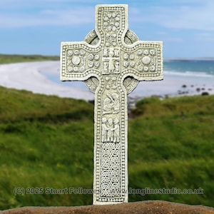 May include: A white stone Celtic cross with intricate carvings and a knotwork design. The cross is standing on a brown surface with a beach and ocean in the background.