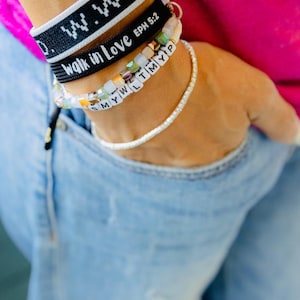 May include: A person's wrist with several bracelets. The bracelets are black with white text, one says "Walk in Love Eph 5:2", and one has white letter beads that spell "SMYWWLLTMYP".