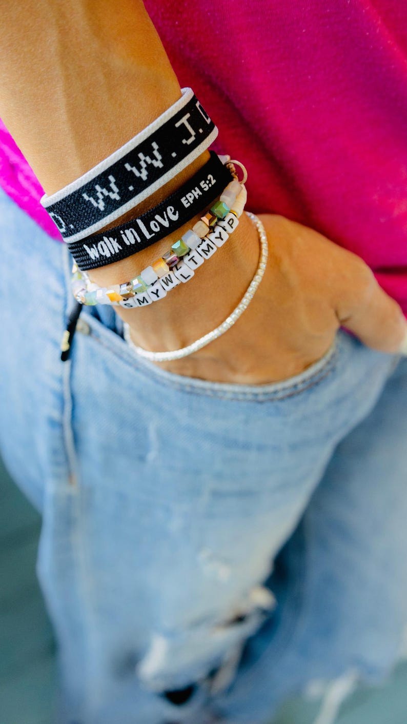 May include: A person's wrist with several bracelets. The bracelets are black with white text, one says "Walk in Love Eph 5:2", and one has white letter beads that spell "SMYWWLLTMYP".
