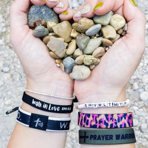 May include: A person's hands holding a heart shape of small grey rocks. The person is wearing several bracelets, including one that says "walk in love Eph 5:2" and another that says "prayer warrior".