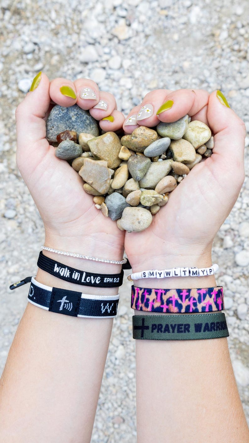 May include: A person's hands holding a heart shape of small grey rocks. The person is wearing several bracelets, including one that says "walk in love Eph 5:2" and another that says "prayer warrior".
